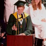 Students wearing graduation caps and gowns. Student speaks at podium.