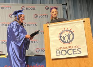 Student and adult at podium. Capital Region BOCES sign. Student in cap and gown holding certificate.