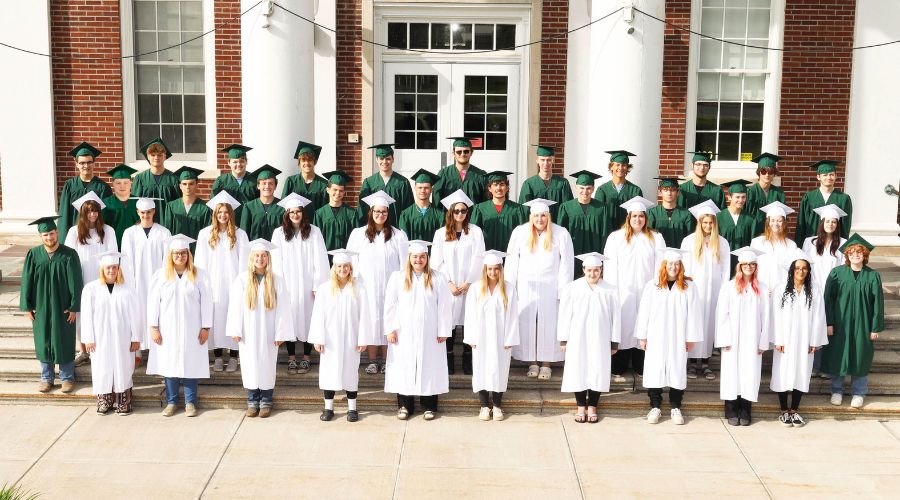 Class of 2024 standing on front steps of school. They are wearing caps and gowns.