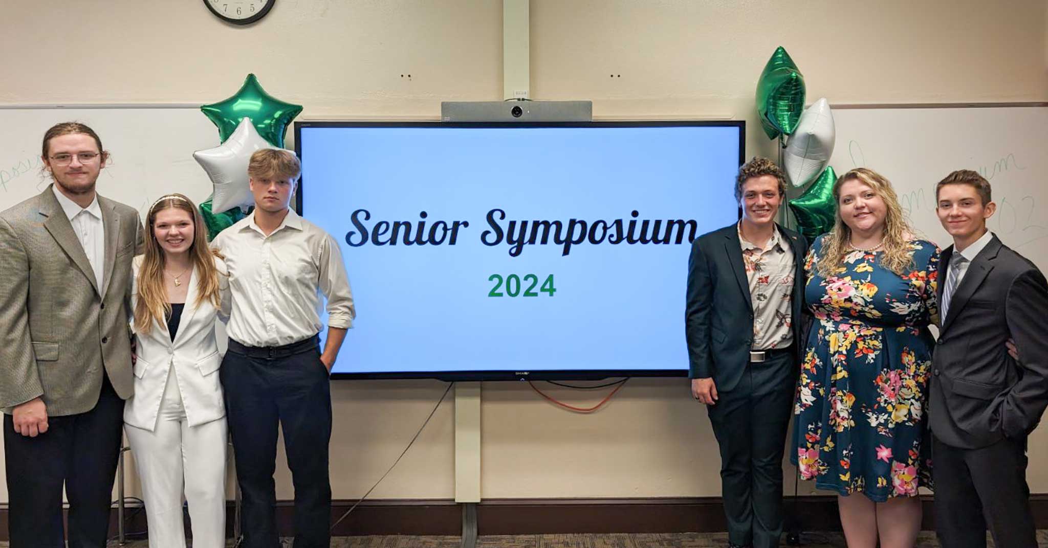 Five students, dressed in professional attire, stand with teacher. Monitor displays, Senior Symposium 2024, green and white balloons in the background.