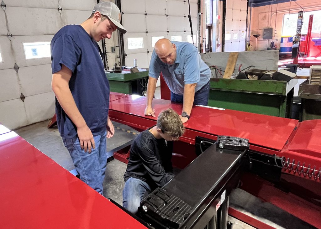 One student and one teacher look on as student works on auto lift.