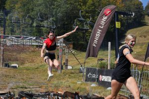 Teen hurdles over a low burning fire at the finish line of a race. Another teen is several steps ahead.