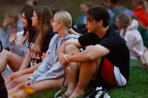 Three teens visible sit on ground with others nearby. Their focus is to the left of camera.