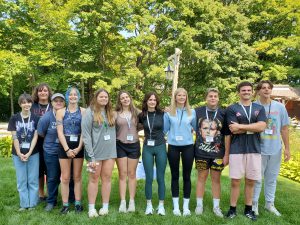 Eleven teens stand in a group, some with arms around each other. They have on lanyards and are dressed for warm weather. They are outside.