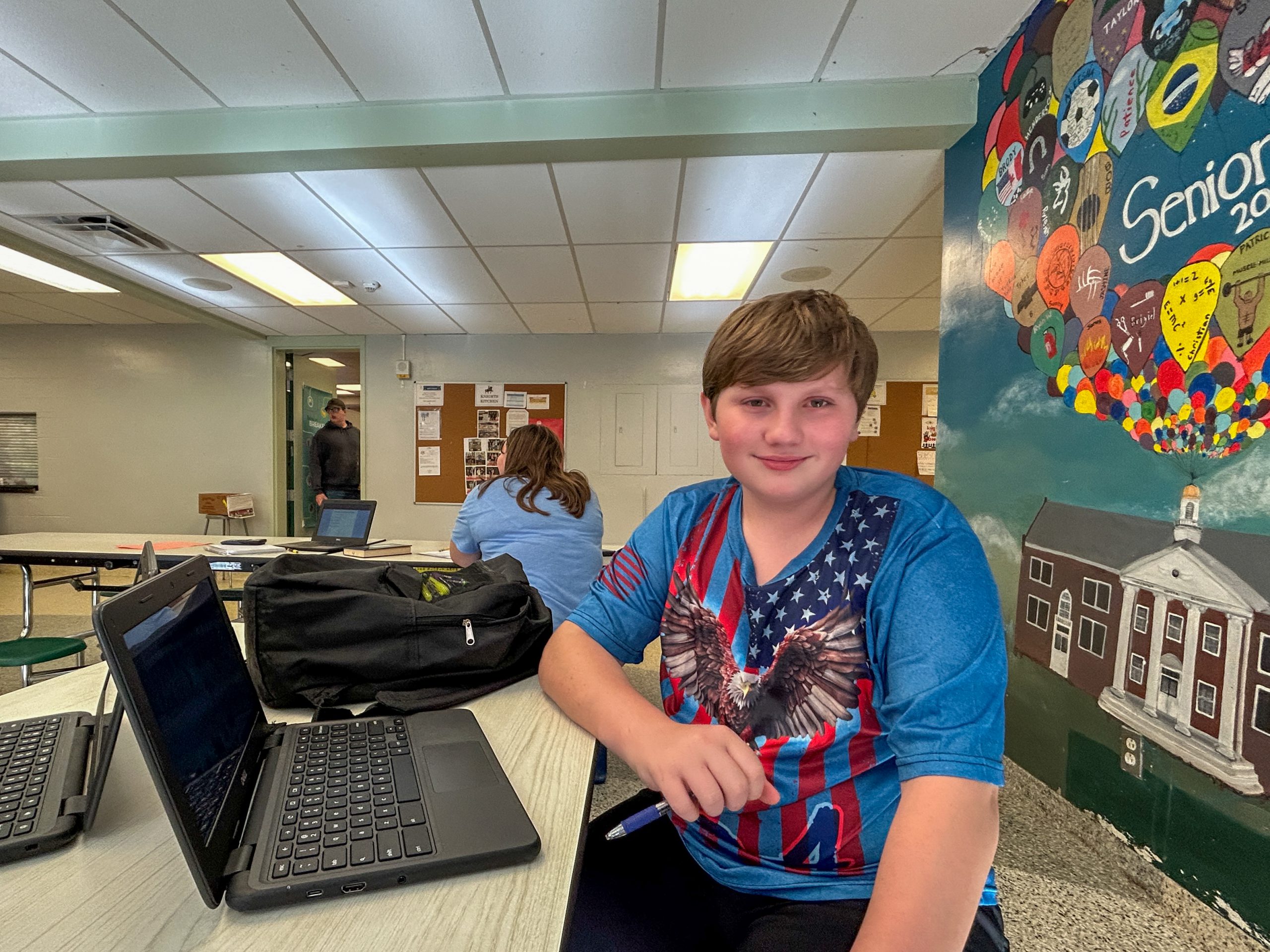 Grade 8 boy sits at table with laptop in front of him. School mural is in background.