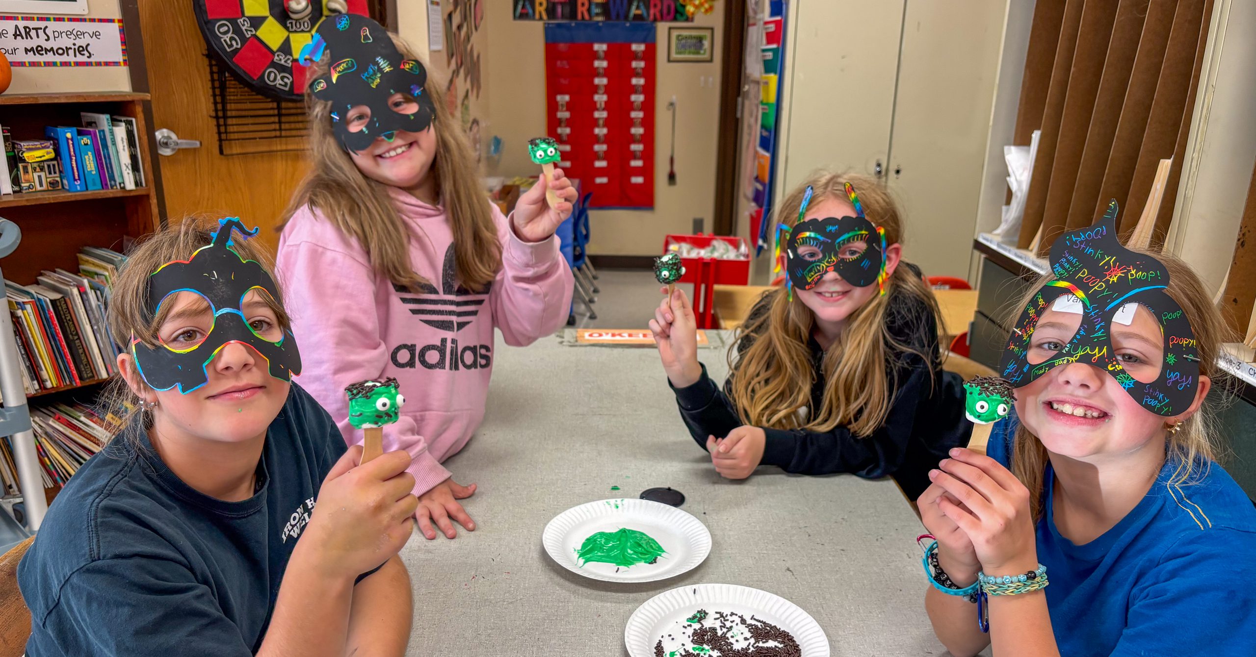 Four students at a table in art room. They are wearing hand-made masks and are holding treats.