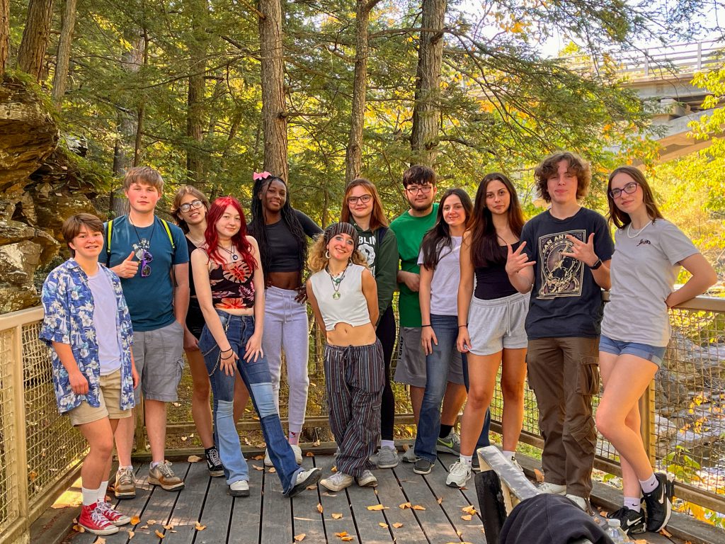 Twelve members of the Class of 2025 stand together on a forest bridge on an early autumn day.