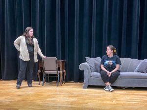 Two students on stage. One is sitting on a couch.