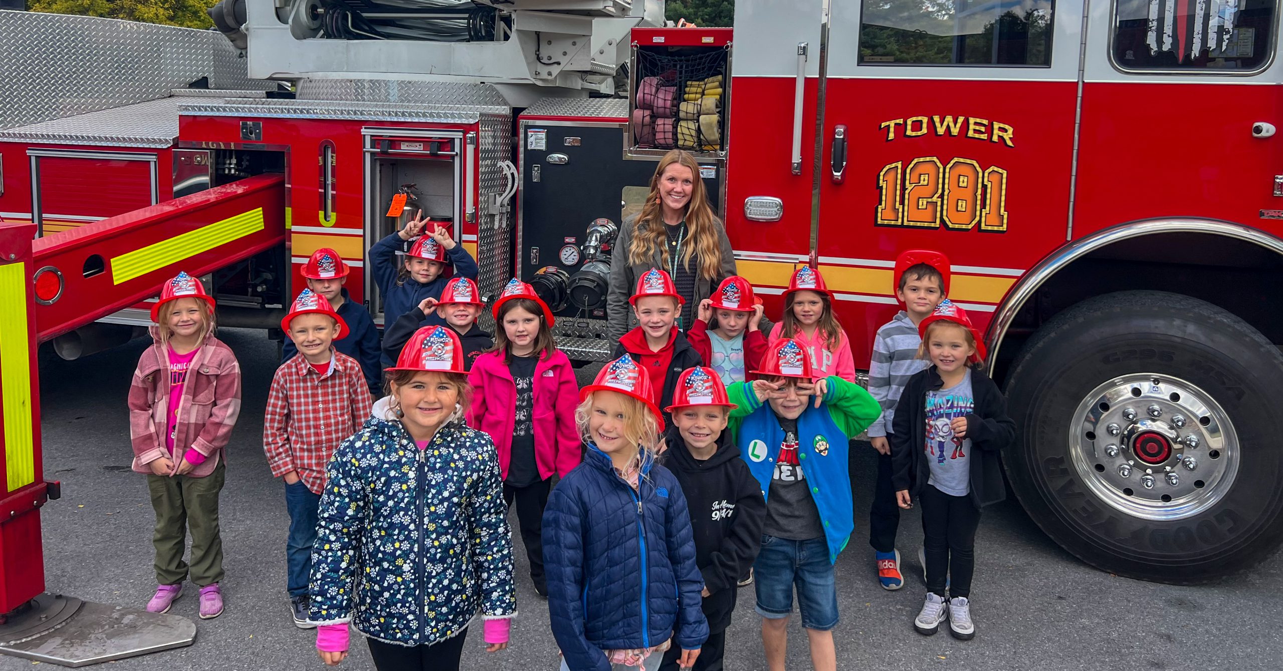 Elementary student stand with teacher next to fire rescue vehicle. Students are wearing plastic fire fighter helmets.