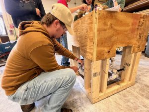 Student squats down to adjust plumbing on a wooden frame. He is wearing a hat.