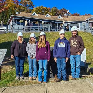 One teacher and four students stand in front of building. Each is wering a hardhat.