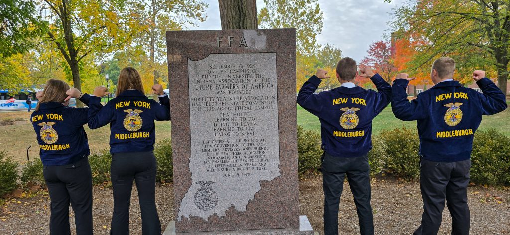 Four students stand next to FFA plaque. Each is facing away from the camera and poinging backward toward their New York Middleburgh FFA jackets.