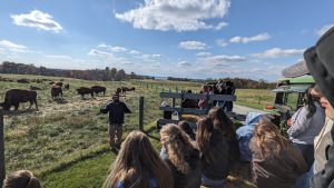 Students sit on tractor-driven wagons listen to rancher. Bison in background.