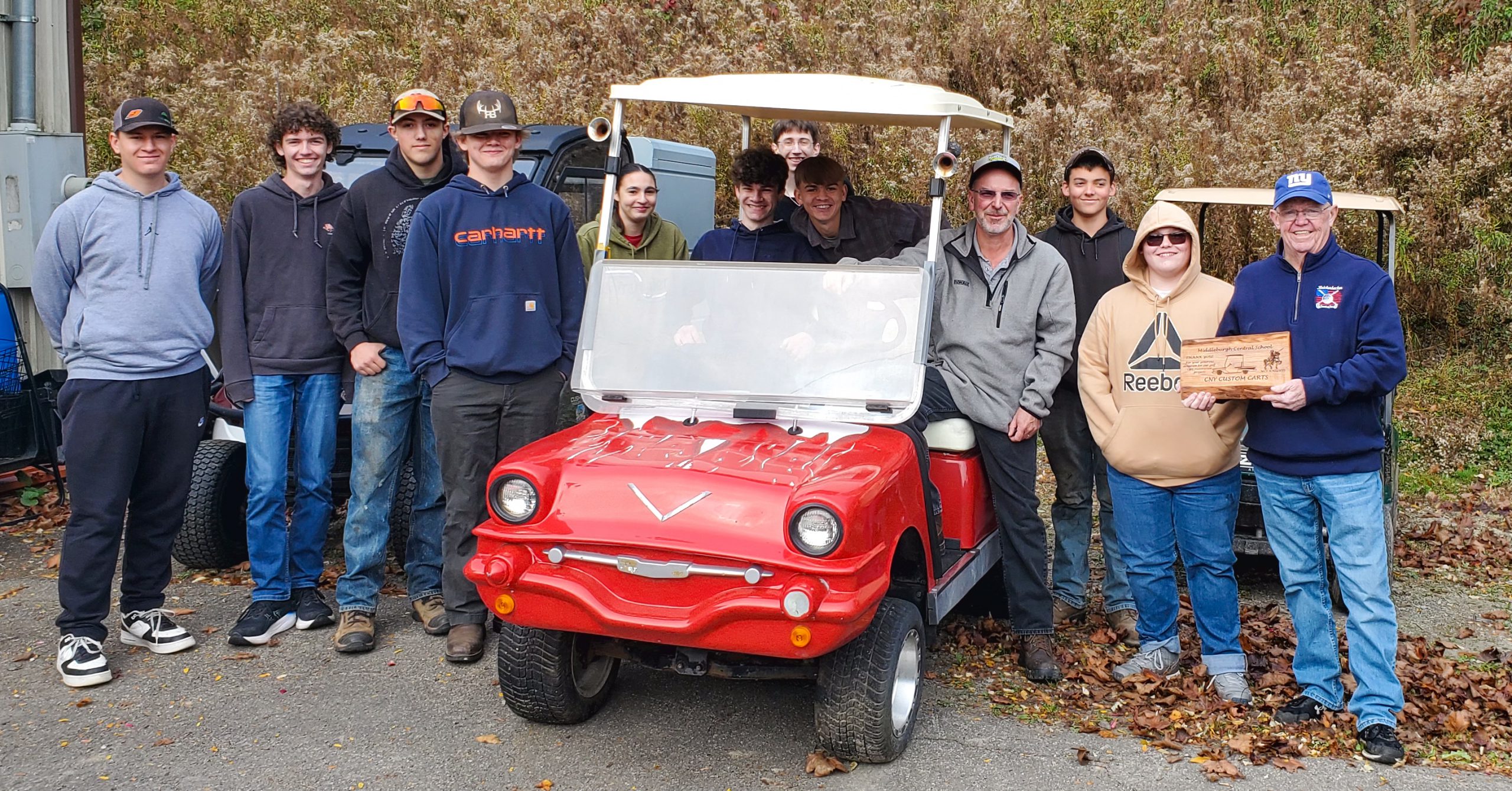 Eleven students and two adults. One adult is holding a wood plaque from CNY Custom Carts. Three students and one adult sit in a cart.