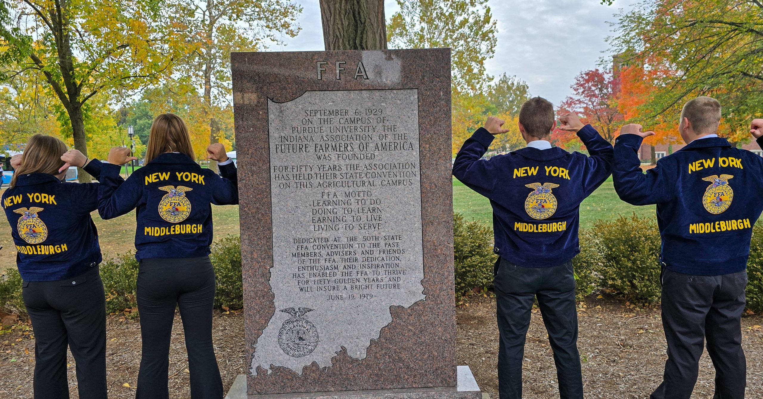 Four students stand beside FFA plaque. Each is facing away from camera and pointing their fingers backward toward their Middleburgh, New York FFA jackets.