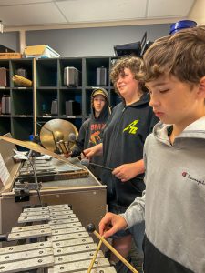 Three teens stand in  percussion section.  One is looking at a xylophone.