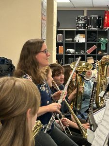 Teacher, holding an instrument, sits among students who are also holding wind instruments.