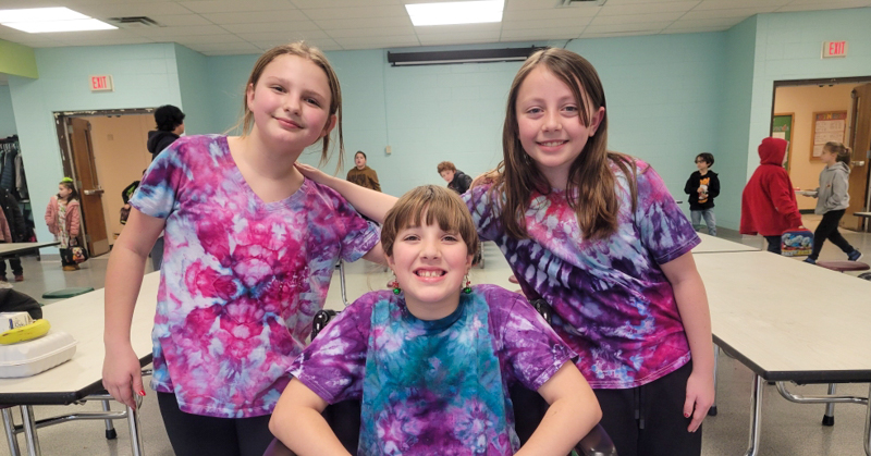 Three students wearing tie-died shirts . One has her arm placed on the shoulder of another student. One is sitting in a wheelchair.