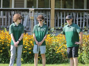 Three members of the varsity golf team stand together with a club in their hands. All are wearing sunglasses.