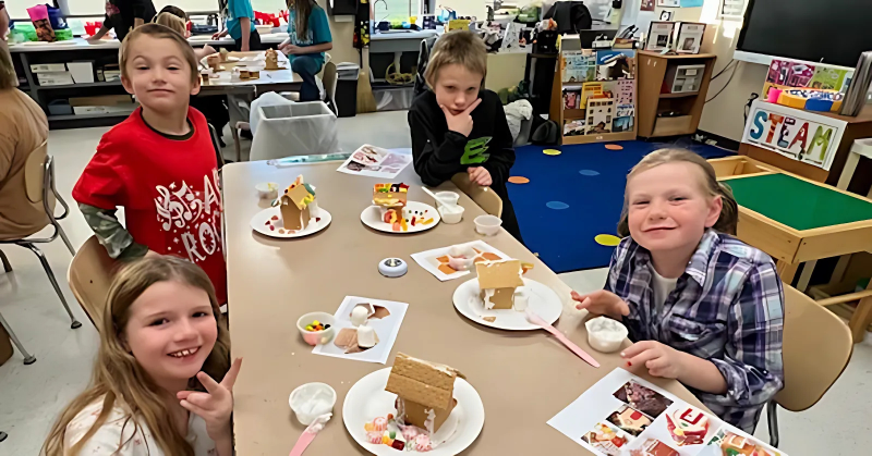 Four elementary students smile and pose at a table. They are making gingerbread houses.