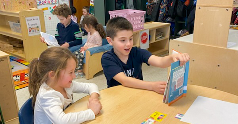 Kindergarten student listens as a third-grade student reads a book to her. Other students do the same in background of classroom.