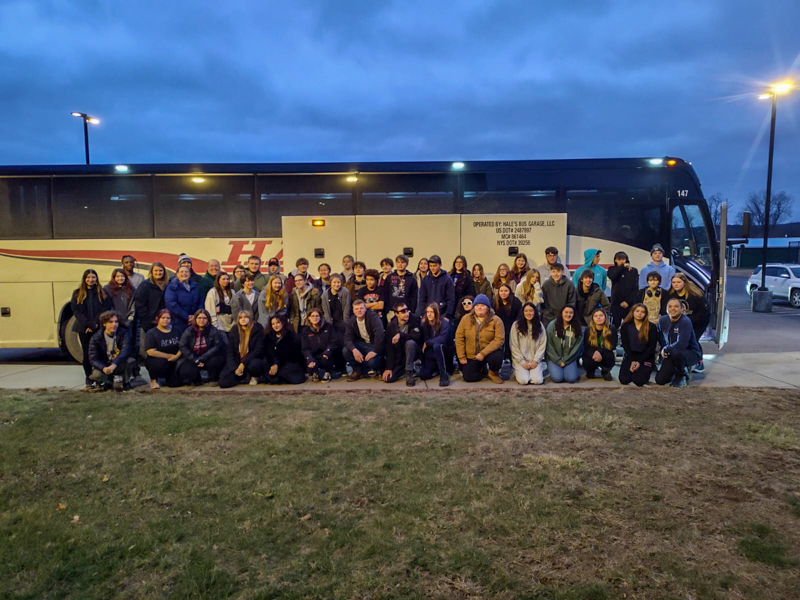 Students and teachers stand outside a tour bus around dawn.