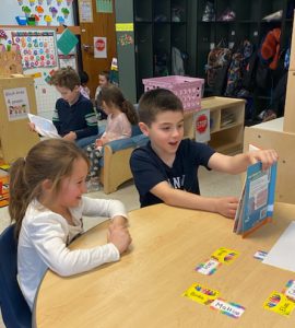 Grade three student next to pre-kindergarten student. Older student is holding a picture book.