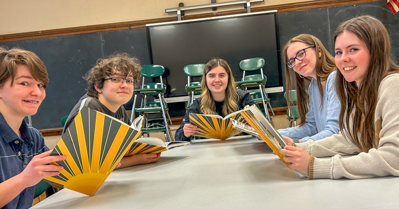 Five students seated at table. Each is holding a book.
