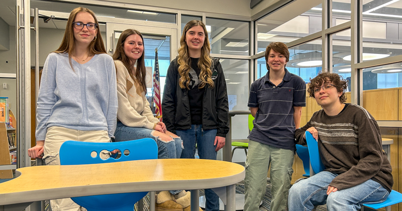 Five high school students standing and sitting in library classroom. Chair and table in foreground.