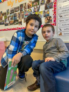 Two boys sit beside each other. One is holding a book.