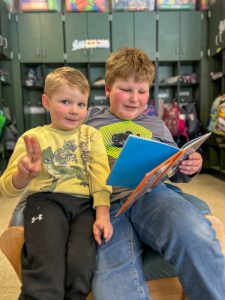 Grade three student next to pre-kindergarten student. Older student is holding a picture book.