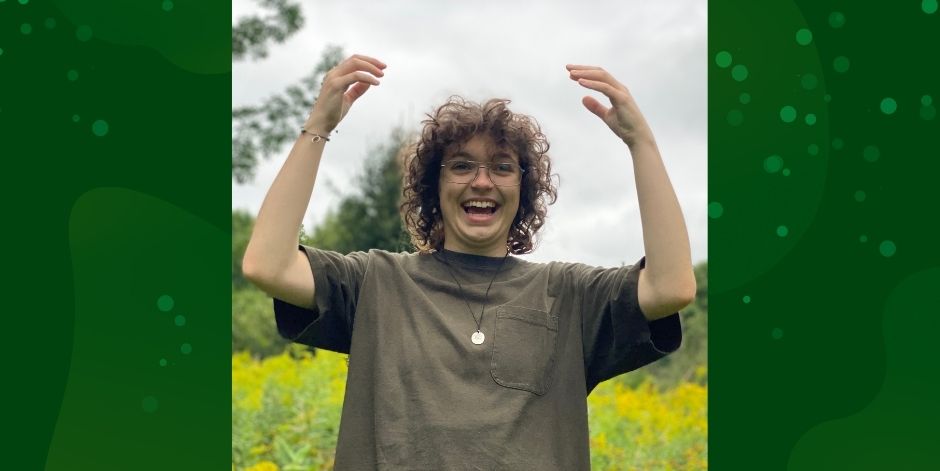 Student raises arms and smiles. They are standing outdoors.