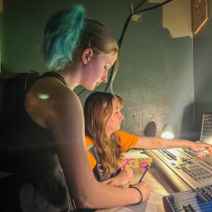 Two teens work in sound and light booth of theater.