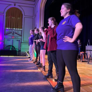 Six teens stand in a row on stage.